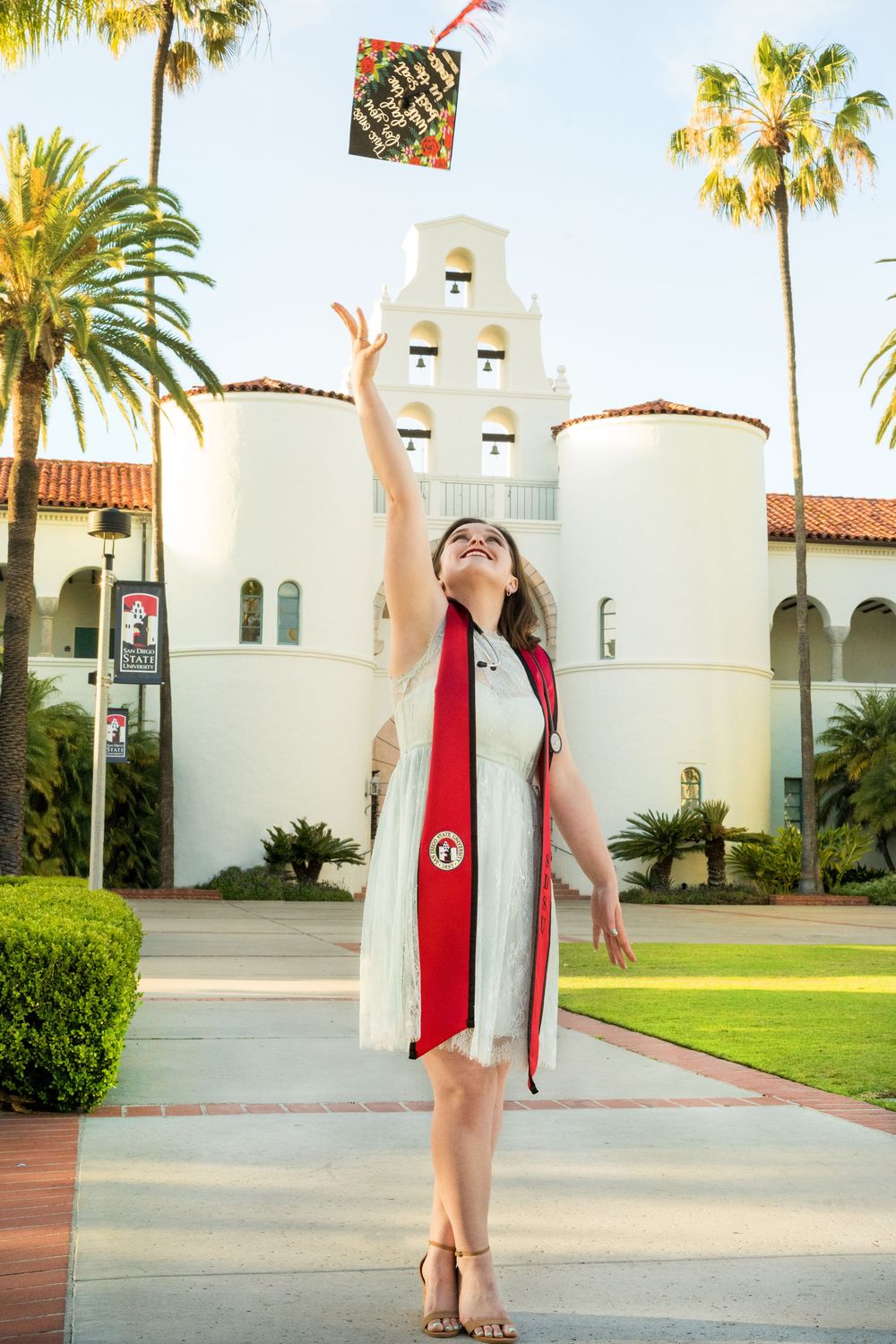 Graduate in white dress and red sash throws graduation cap in front of Spanish-style university building with palm trees.