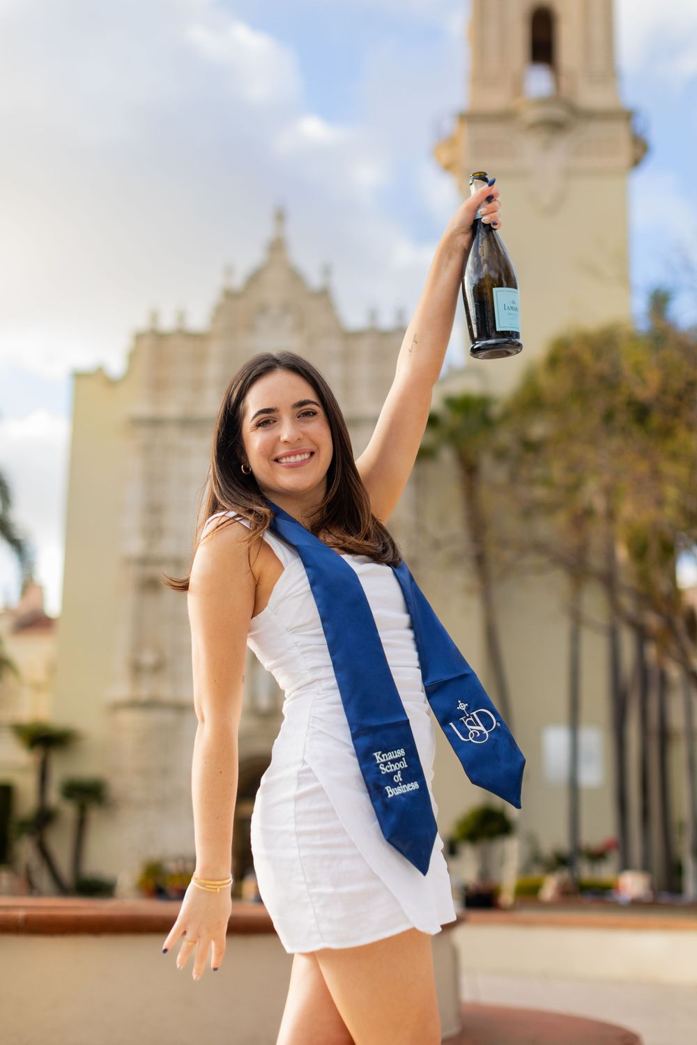 Graduate celebrating in white dress and blue sash holding up champagne bottle in front of historic church building.