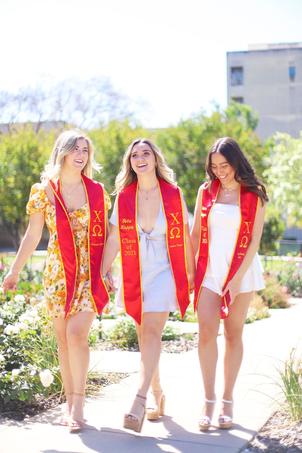 Three sorority members wearing red graduation stoles and summer dresses walk together outdoors while laughing.