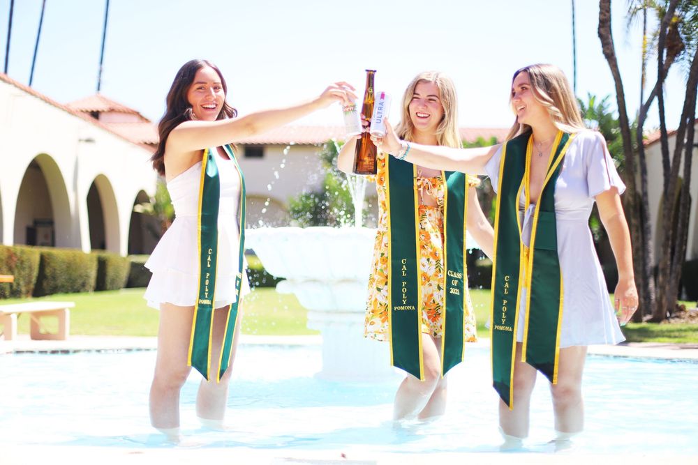 Three graduates wearing green and gold graduation stoles celebrate joyfully outdoors on sunny campus grounds.