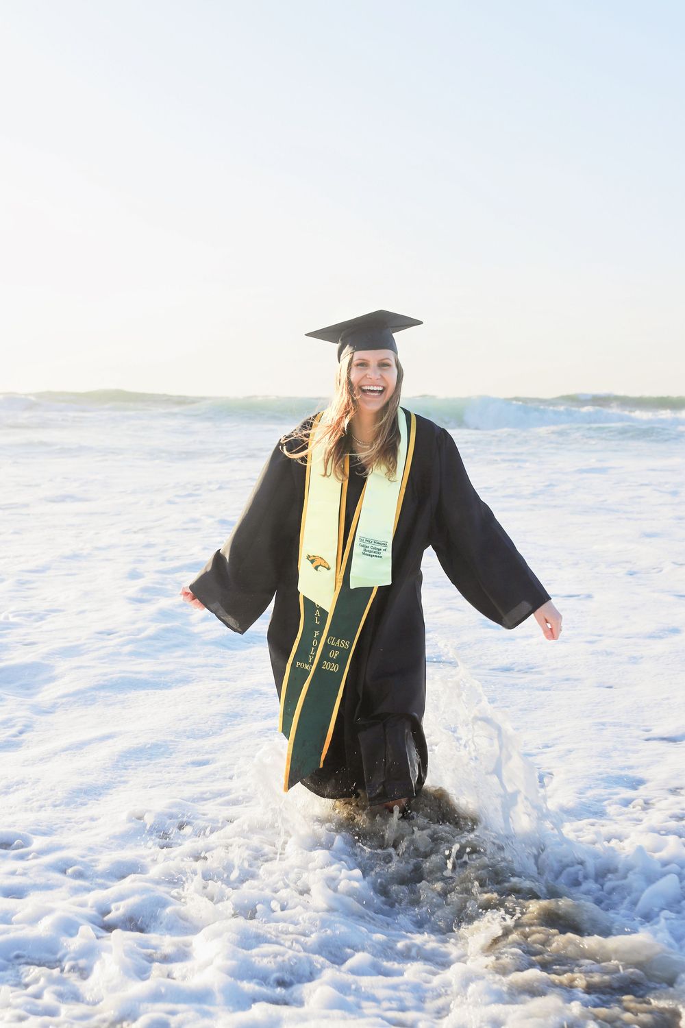 Graduate in black cap and gown stands joyfully in ocean waves at beach during sunset celebration photoshoot.