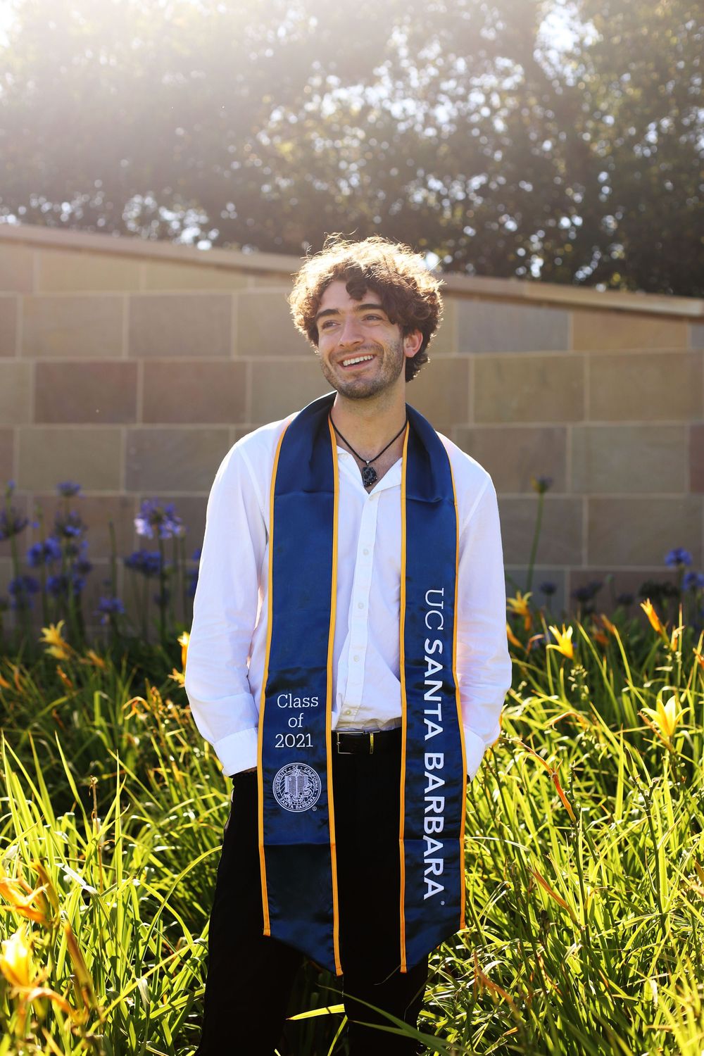Graduate wearing a blue and gold academic stole stands smiling in sunlit garden with purple flowers and stone wall background.