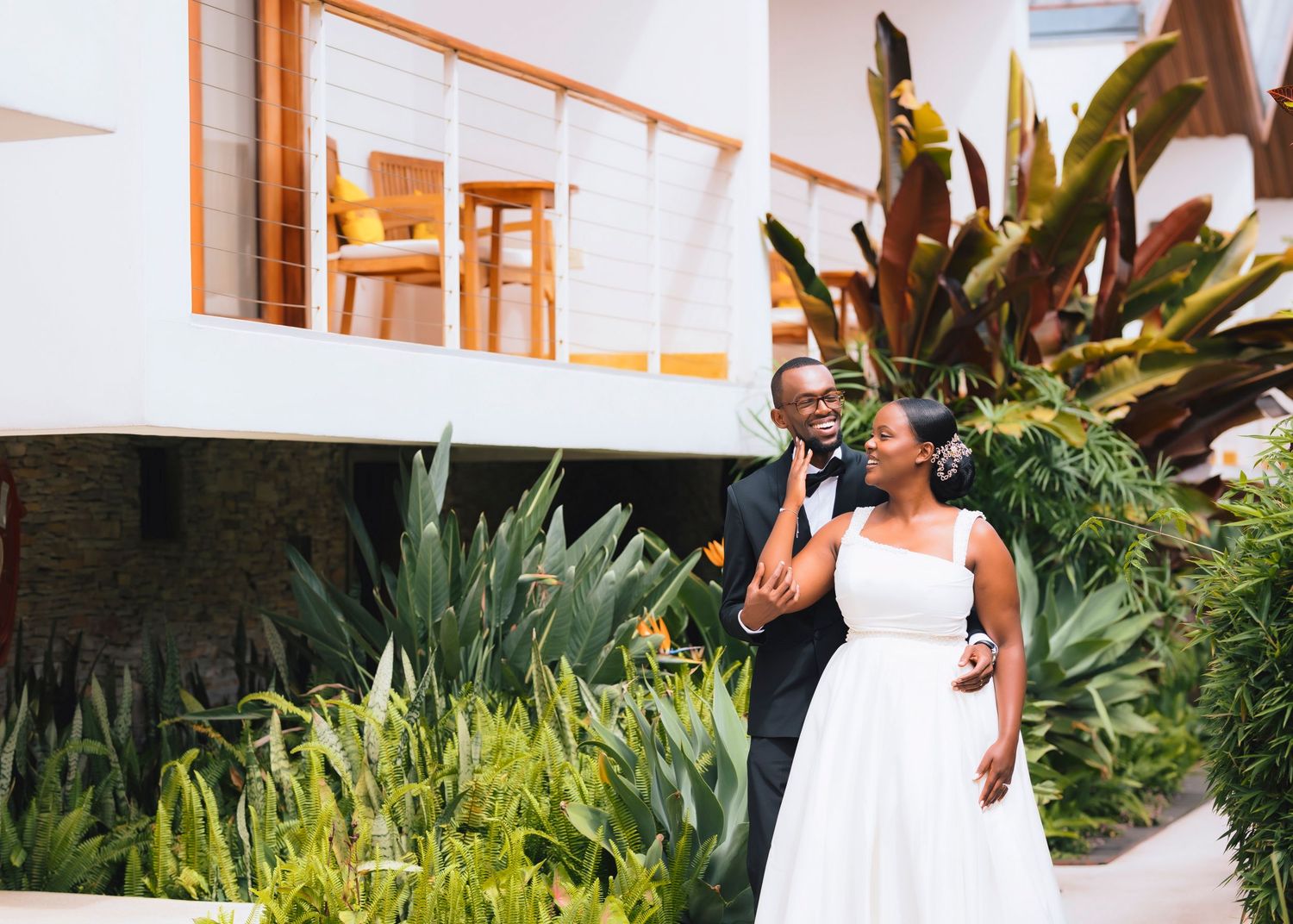 Someone in a flowing white dress stands in front of tropical plants and modern architecture on a sunny day.