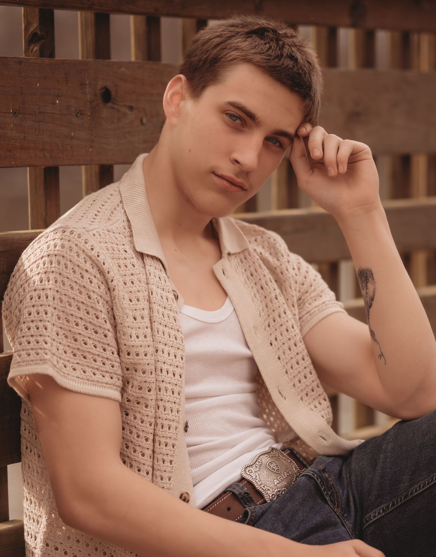 Young model wearing a cream crochet cardigan and white tank top poses against wooden fence in warm lighting.