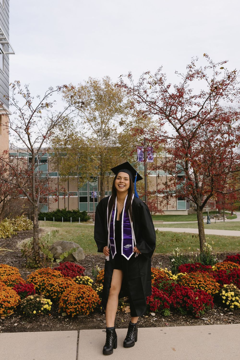 Graduate in cap and gown poses near colorful autumn flowers and trees on a college campus.