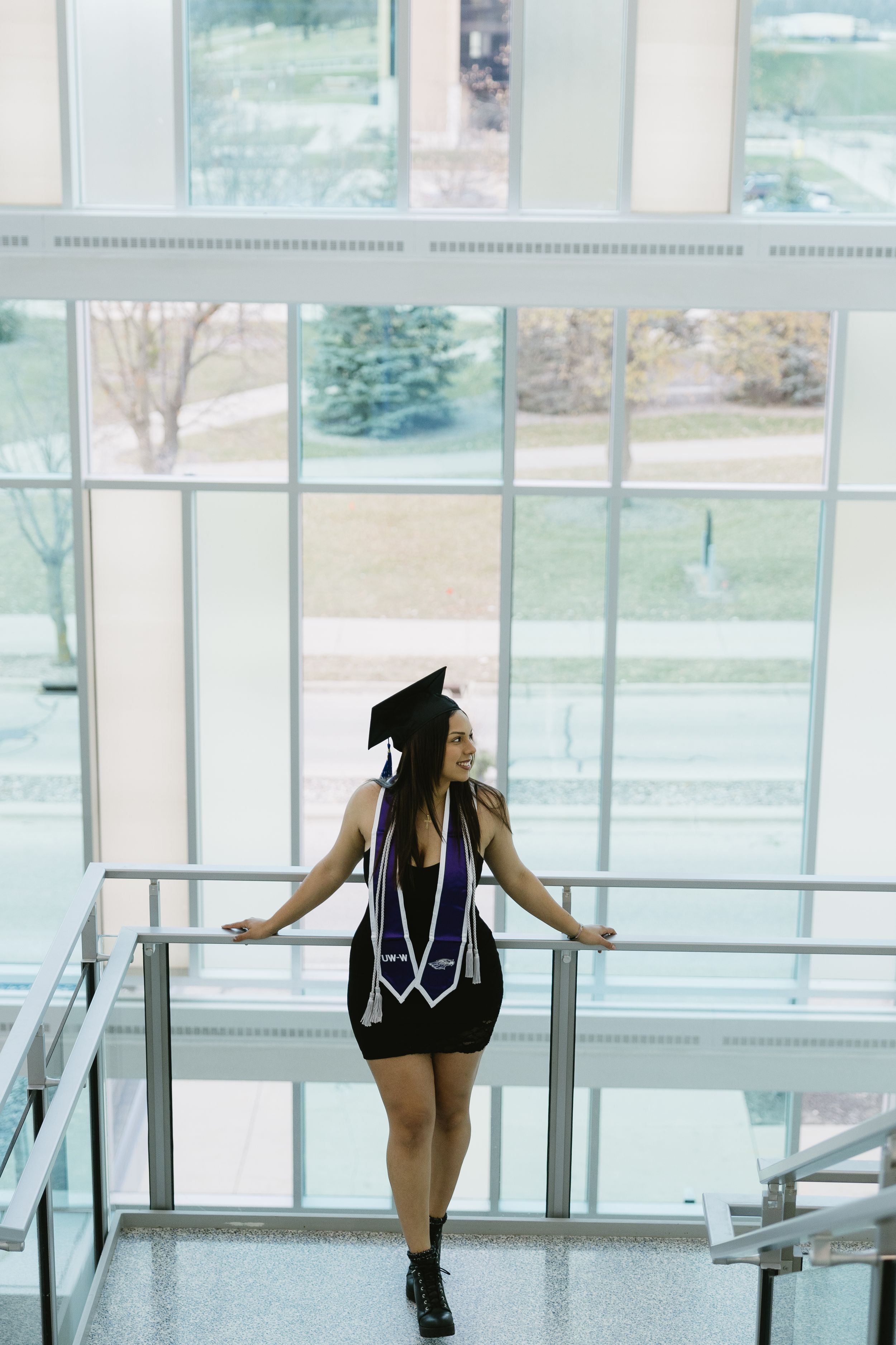 Graduate in cap and gown standing near large windows overlooking a sunny campus lawn.