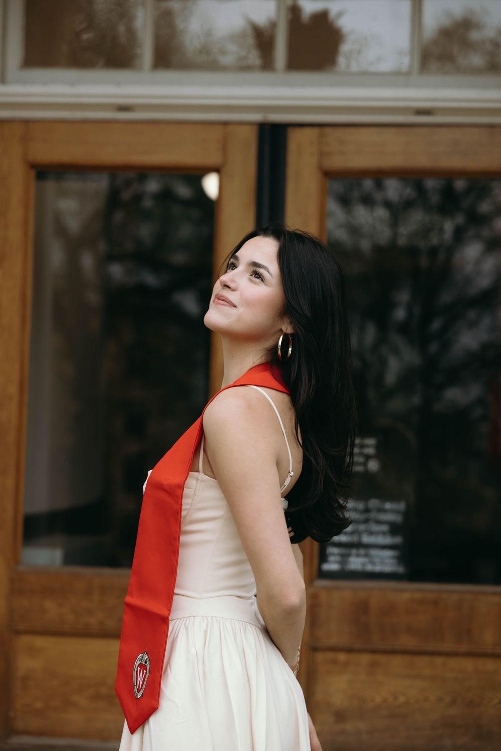 Woman in white dress and red scarf looking upward against wooden storefront with windows.