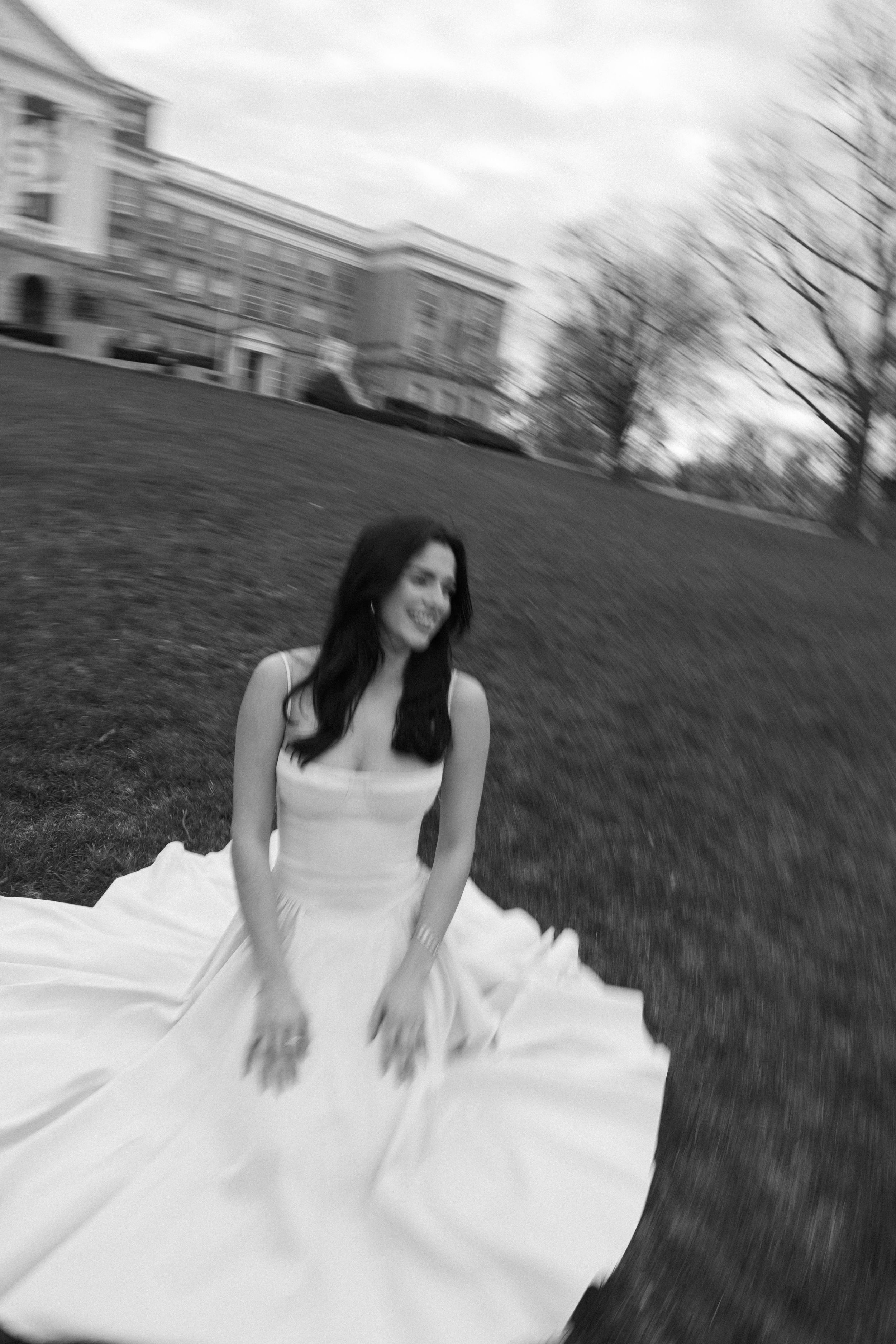 Black and white photo of someone in a flowing white wedding dress sitting on a grassy hill near buildings.