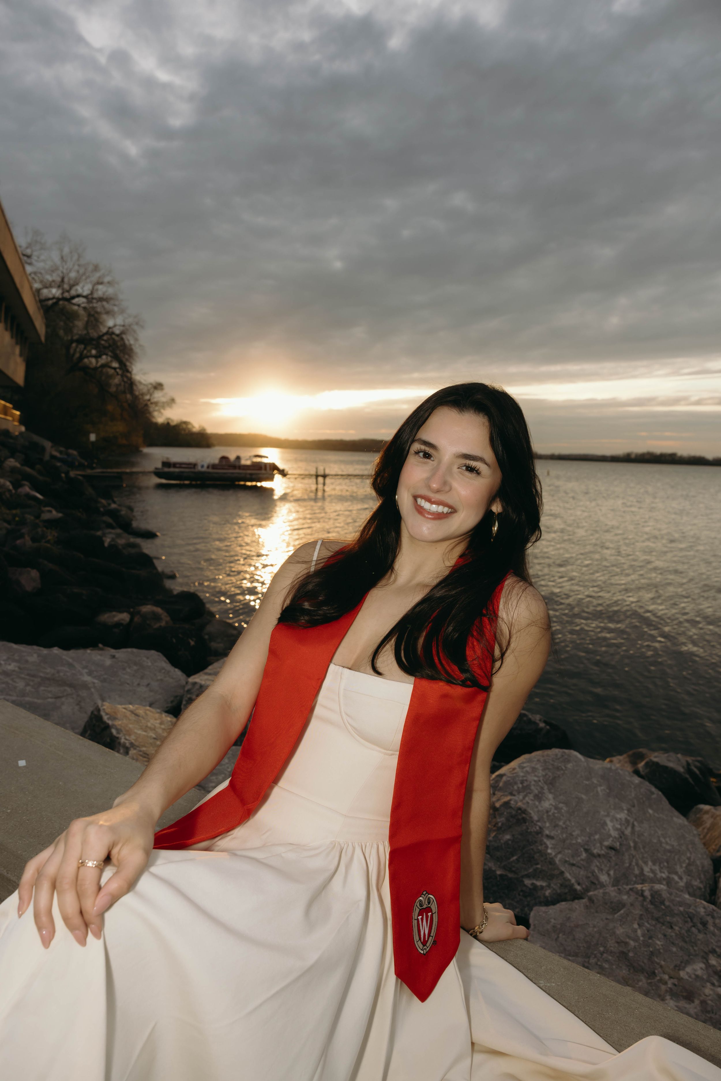 A scenic sunset portrait by the water featuring a white dress and red graduation stole against a cloudy evening sky.