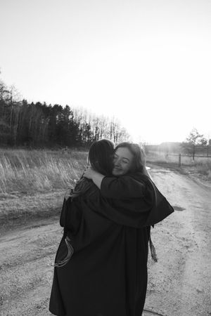 Two people in graduation robes share a heartfelt hug on a dirt road with trees and sunlight in the background.