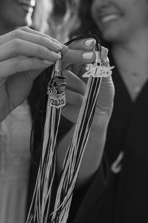 A close-up black and white photo showing hands holding graduation tassels with 2025 charms.