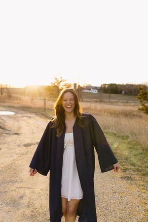 Person wearing black graduation gown and white dress stands on dirt path during golden sunset with countryside in background.