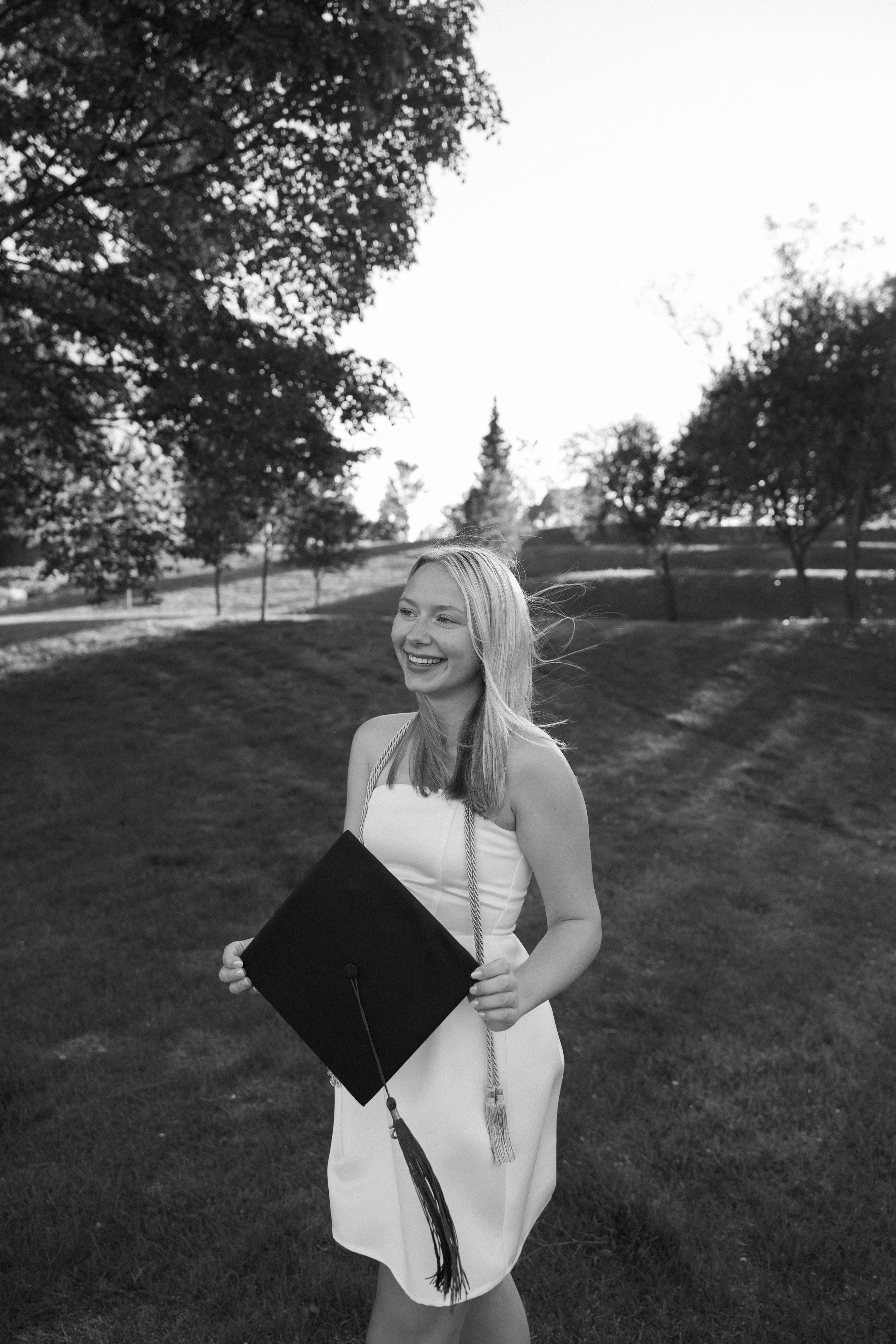 A graduate in a white dress holds a black diploma folder while standing on a grassy hillside in black and white.