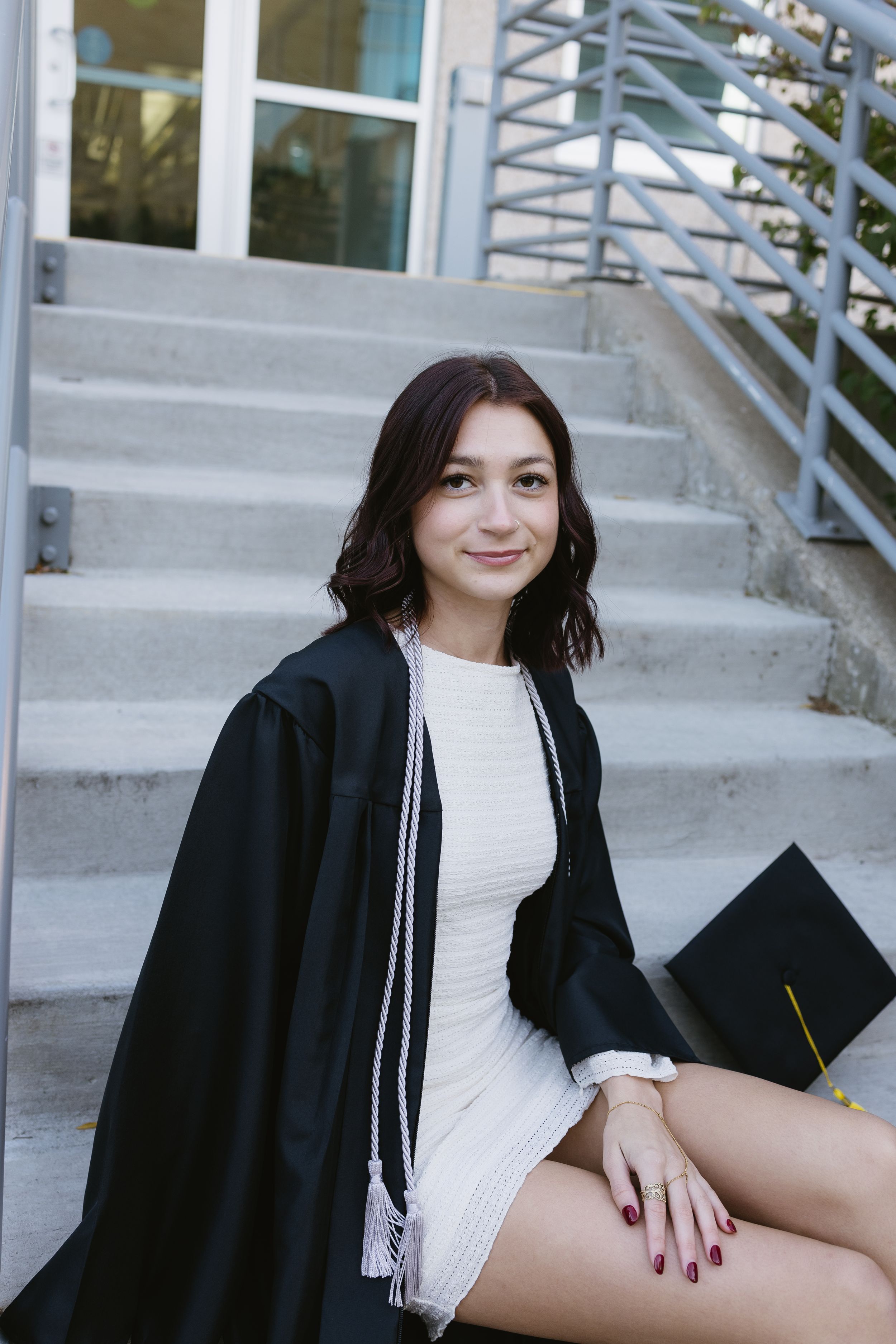 Graduate wearing black cap and gown with white dress sits on concrete stairs during graduation photoshoot.