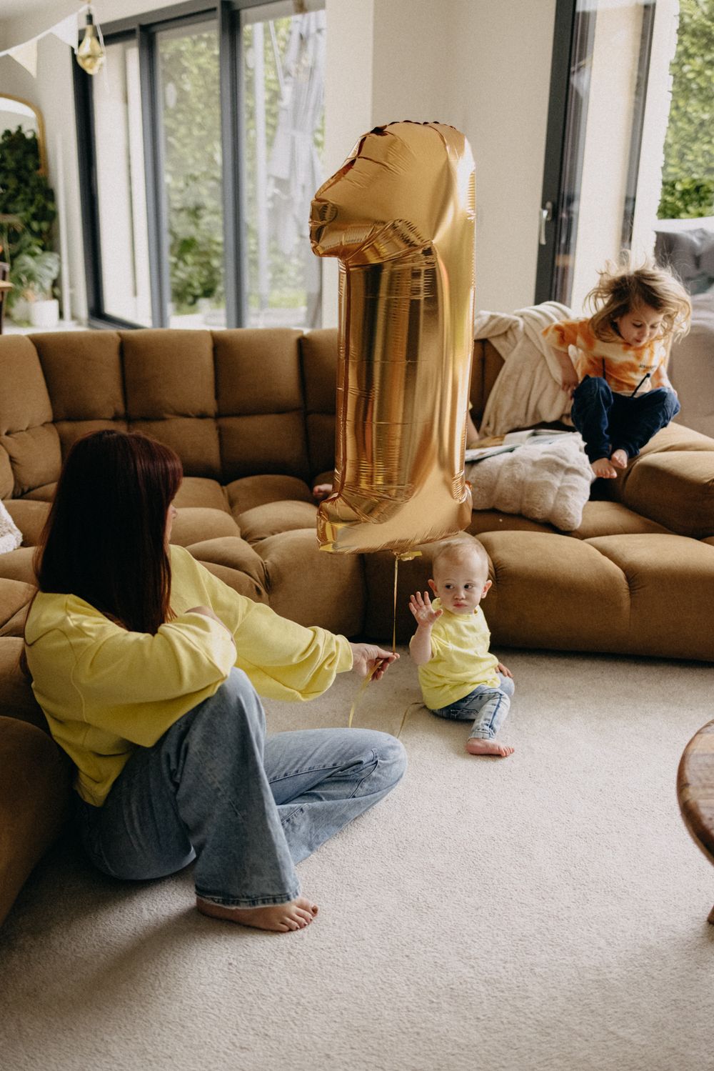 A family plays with a large gold number 1 balloon in their living room with a brown couch and carpeted floor.