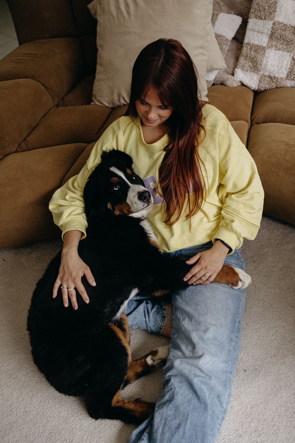 A person in a yellow sweatshirt sits on a brown couch while cuddling with a large black and white dog.