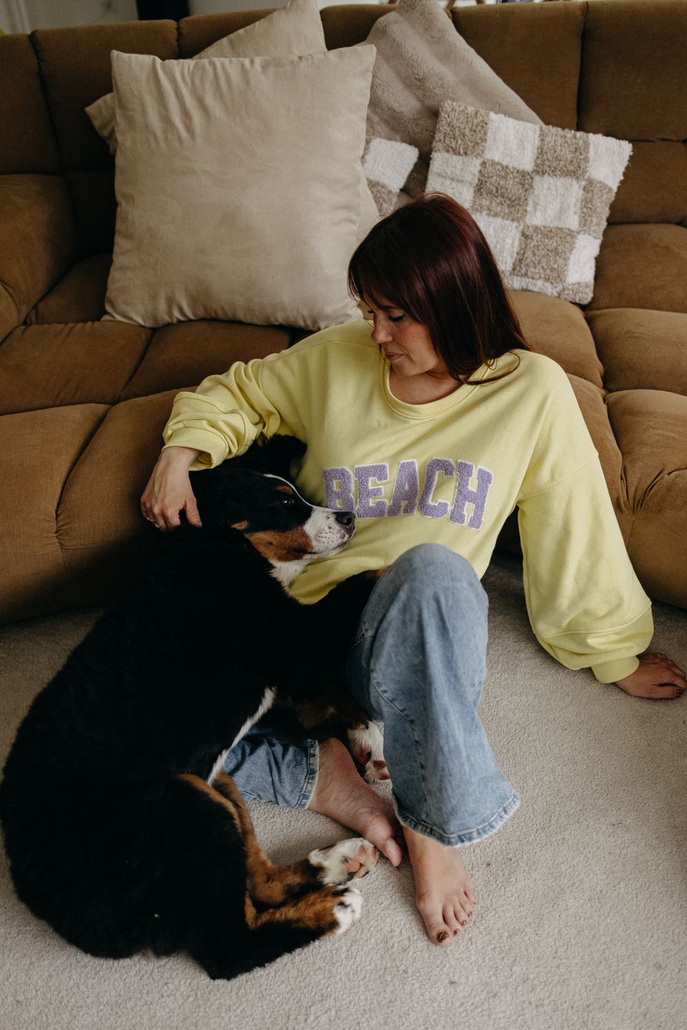 A person wearing a yellow sweatshirt relaxes on a brown couch with their dog on the carpet.