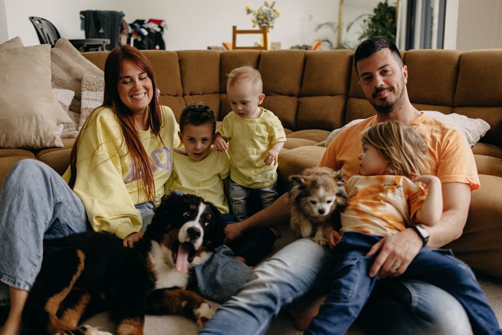 A family relaxes together on a brown couch with their two pets, a dog and cat, all wearing coordinating yellow and orange outfits.
