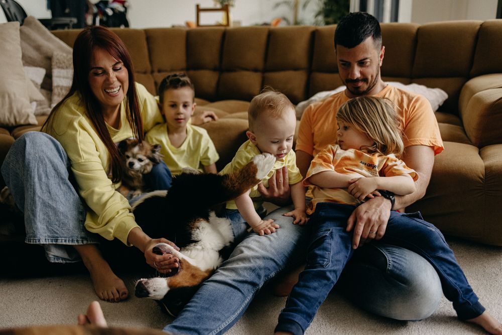 A family relaxes together on a couch with their children and pet dog in a cozy living room setting.