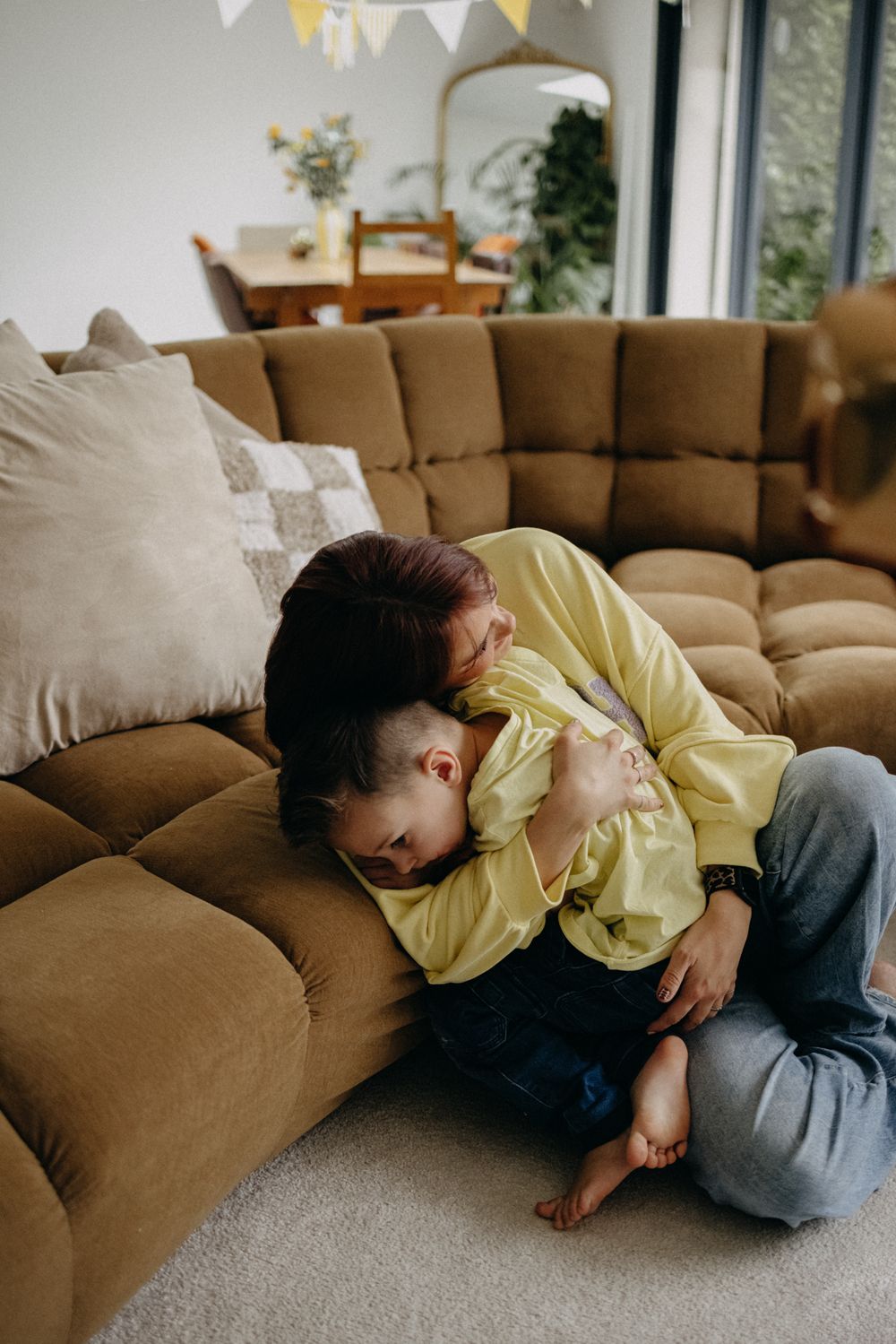 Two people share a tender moment together while resting on a brown sectional sofa beneath decorative bunting.