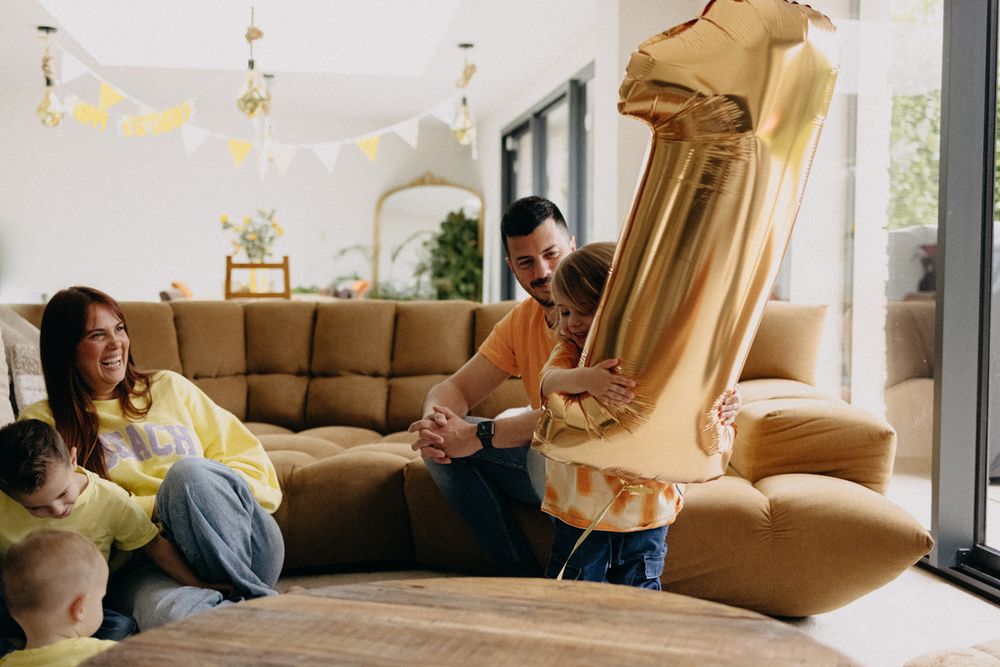 Two friends relaxing on a brown sectional couch while playing with a large golden number one balloon in a living room.