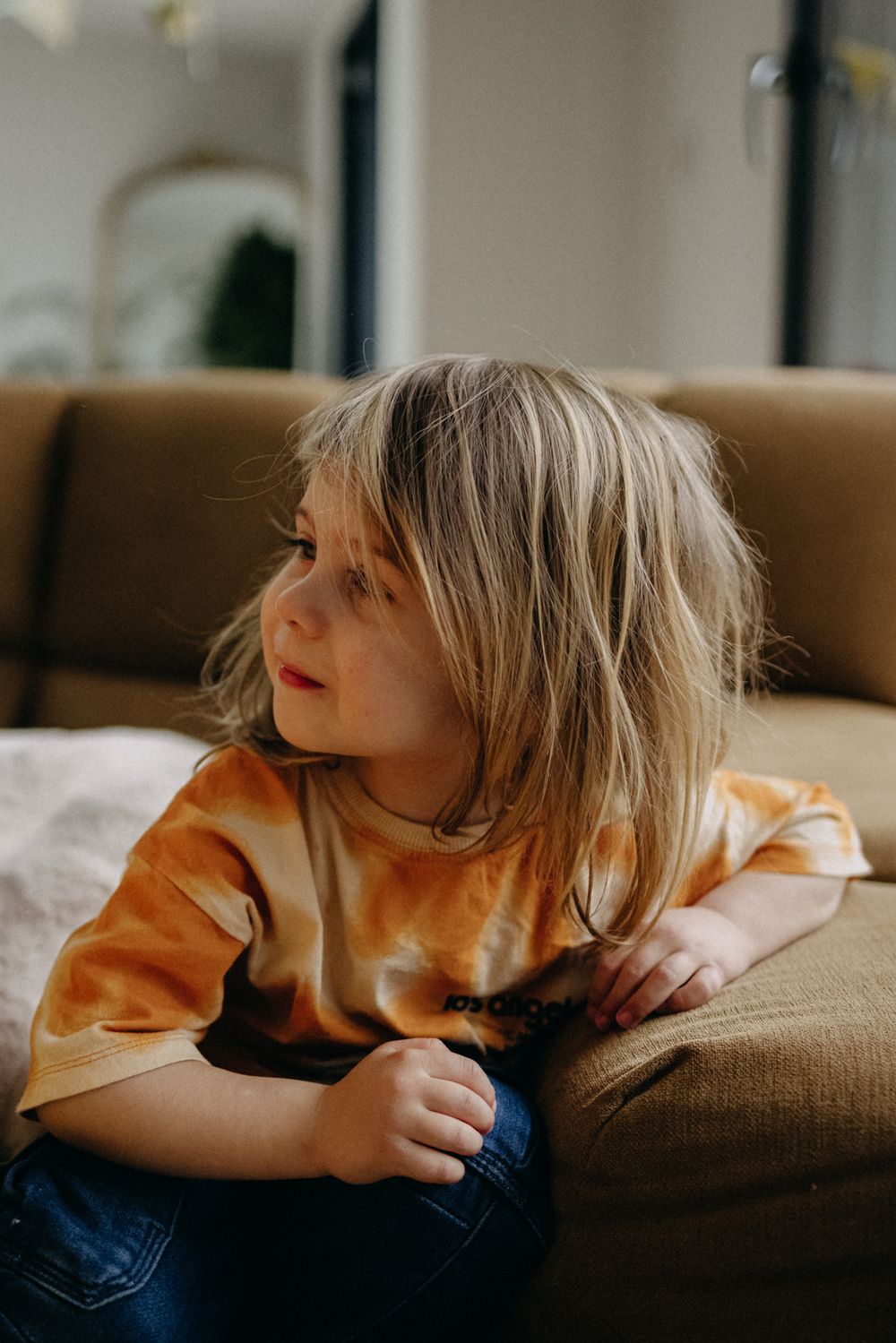 A child in an orange sweater and jeans sits thoughtfully on a brown couch while looking to the side.
