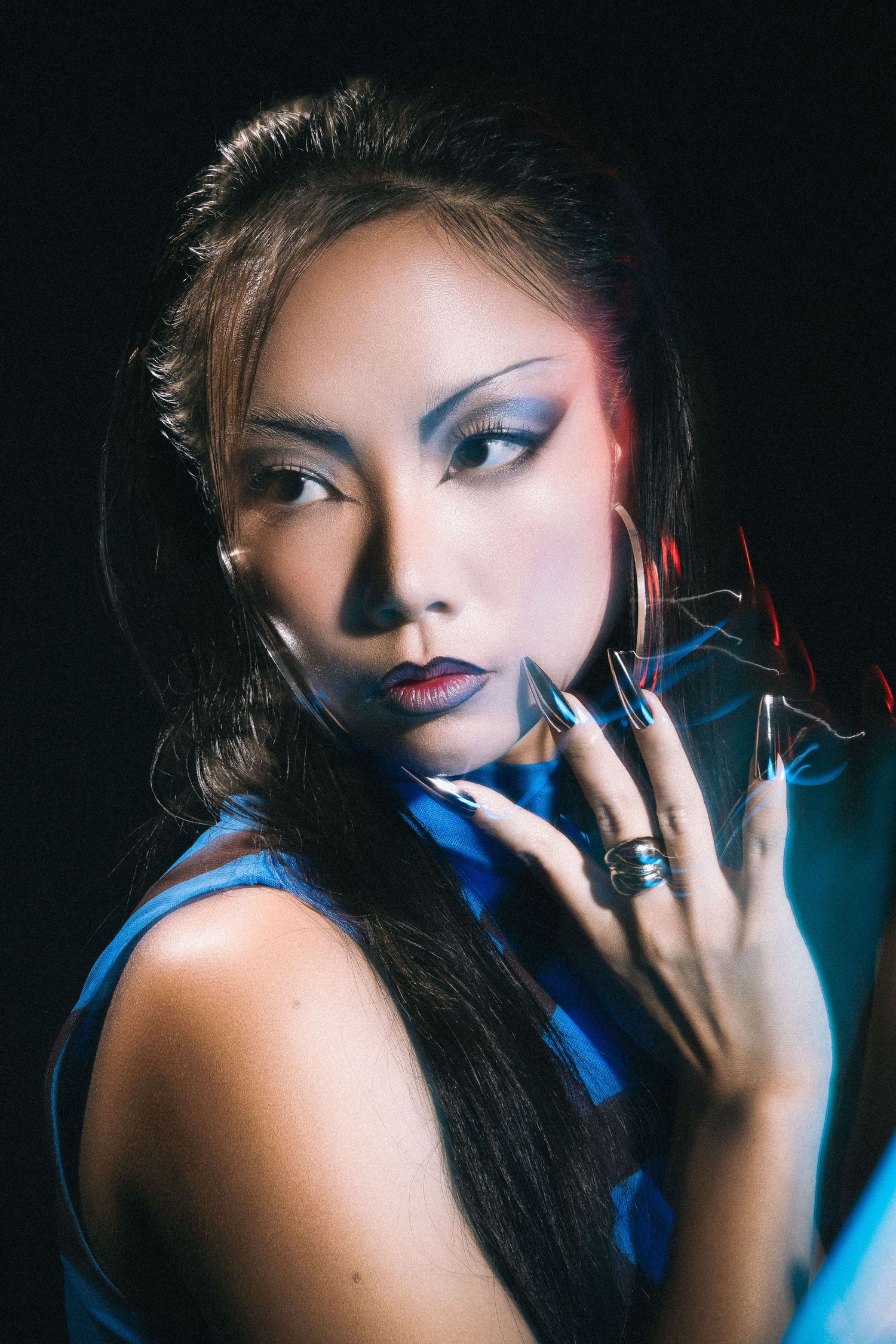 Dramatic beauty portrait with bold blue lighting and creative makeup against dark background.