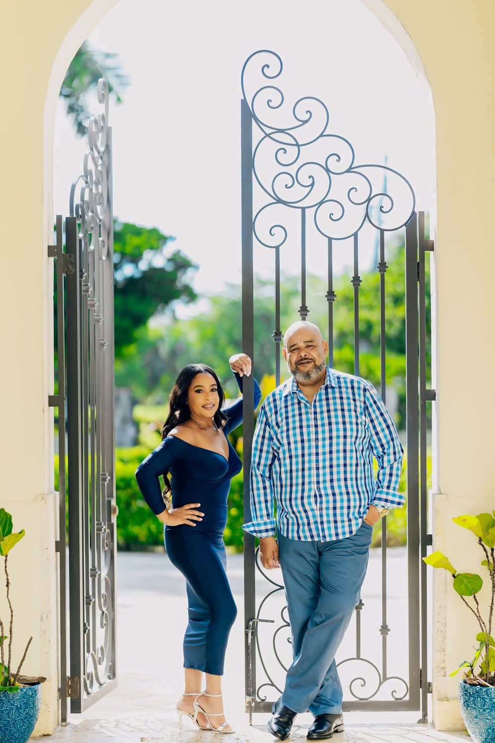 A couple stands together in a decorative arched entryway with ornate wrought iron scrollwork and lush greenery in the background.