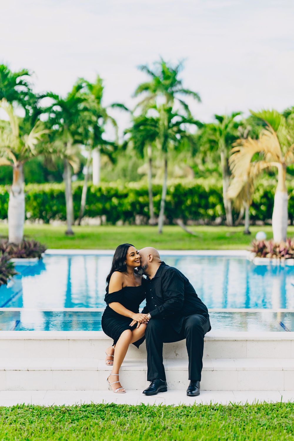 Couple sharing a romantic moment by a luxurious pool surrounded by palm trees at a tropical resort.