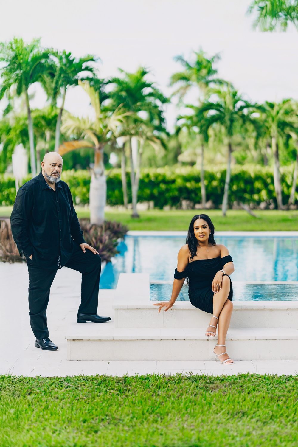 Two people pose by a luxurious swimming pool surrounded by tropical palm trees and landscaped gardens.