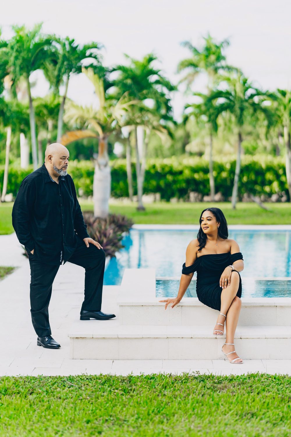 Couple posing for a photo by a swimming pool with palm trees in a tropical setting.