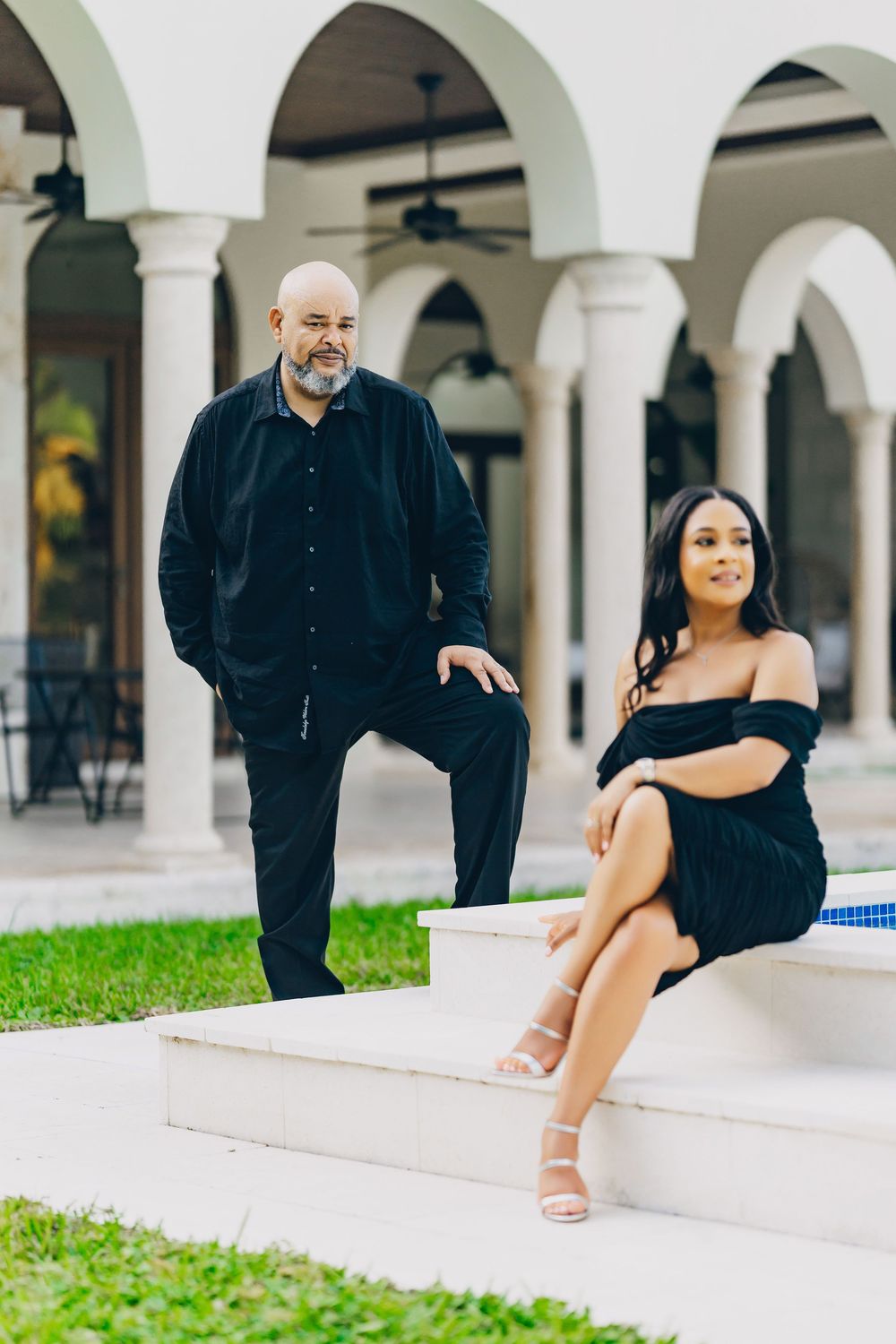 Couple posing together under white arched columns in formal attire.