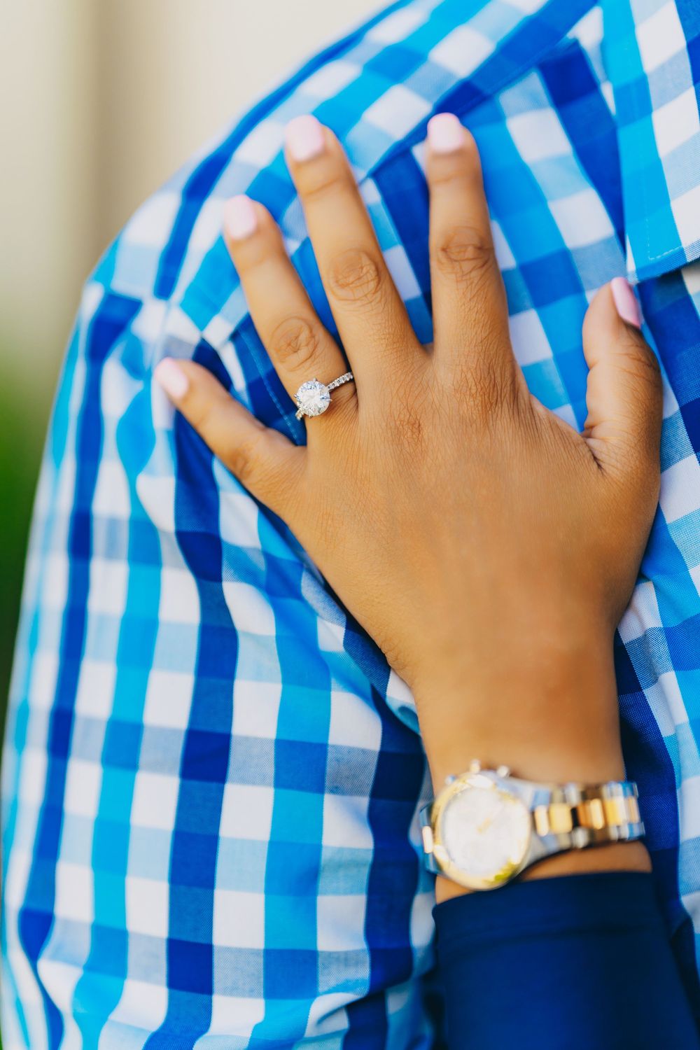 Hand wearing engagement ring and watch rests against blue and white checkered shirt.