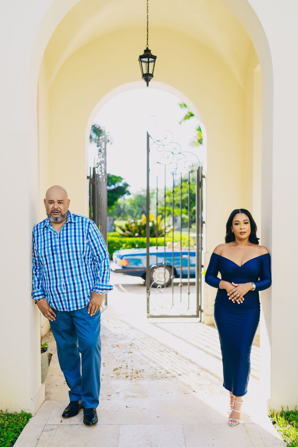 Two people pose in an elegant archway with blue outfits against a cream colored Mediterranean style backdrop and tropical foliage.