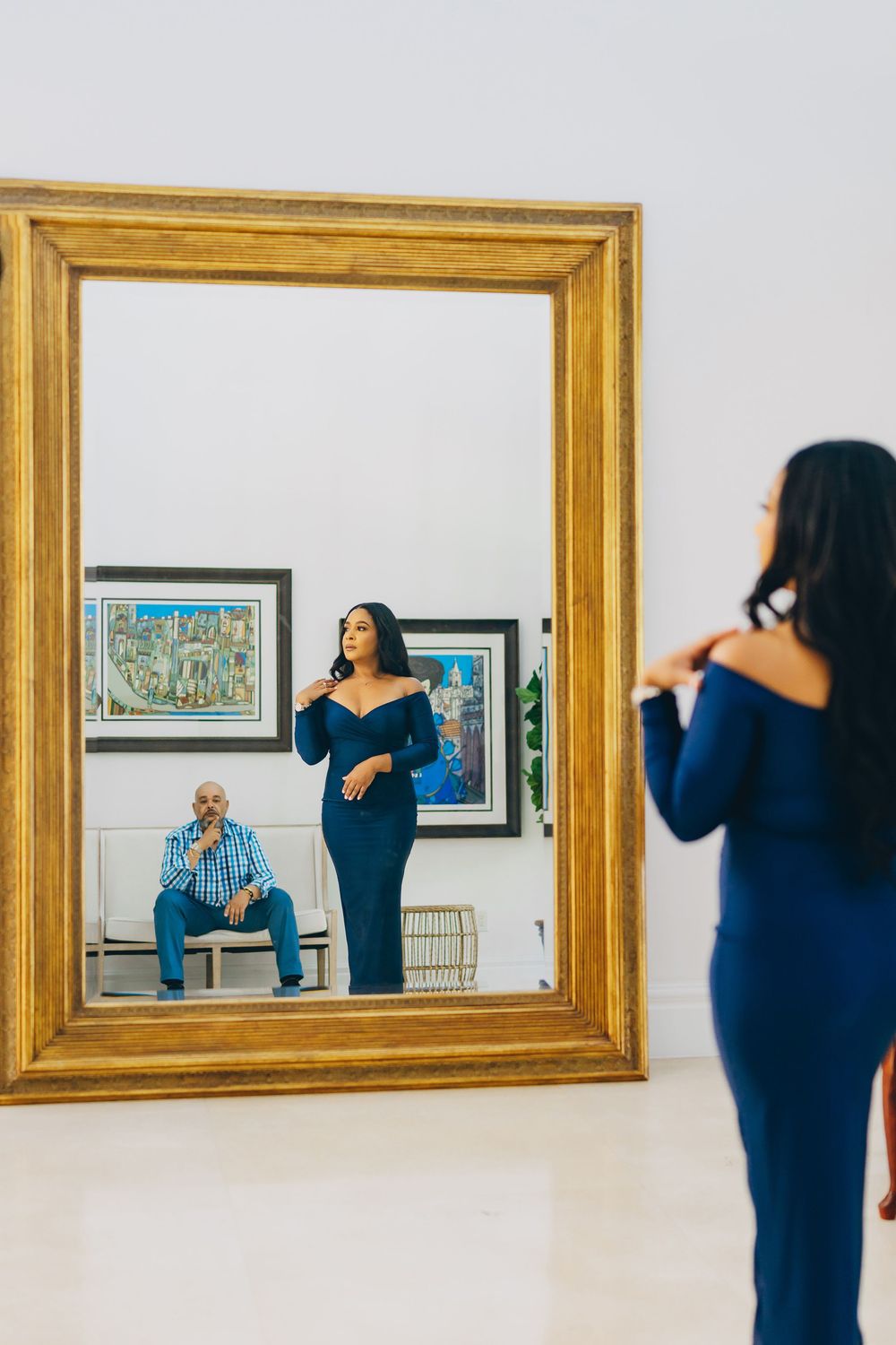 A reflection in a gold-framed mirror shows a woman in a navy dress posing while standing in an art gallery interior.