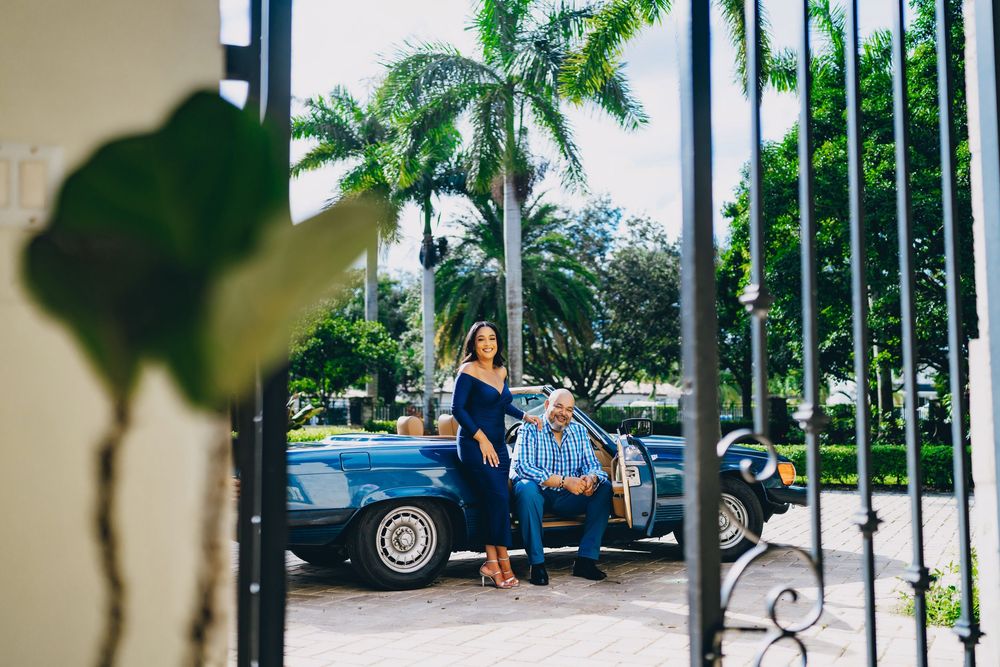 A couple stands beside a vintage blue car under tall palm trees in a tropical setting.