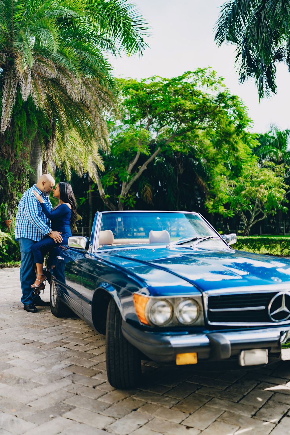A classic blue Mercedes-Benz convertible parked under tropical palm trees on a stone driveway.