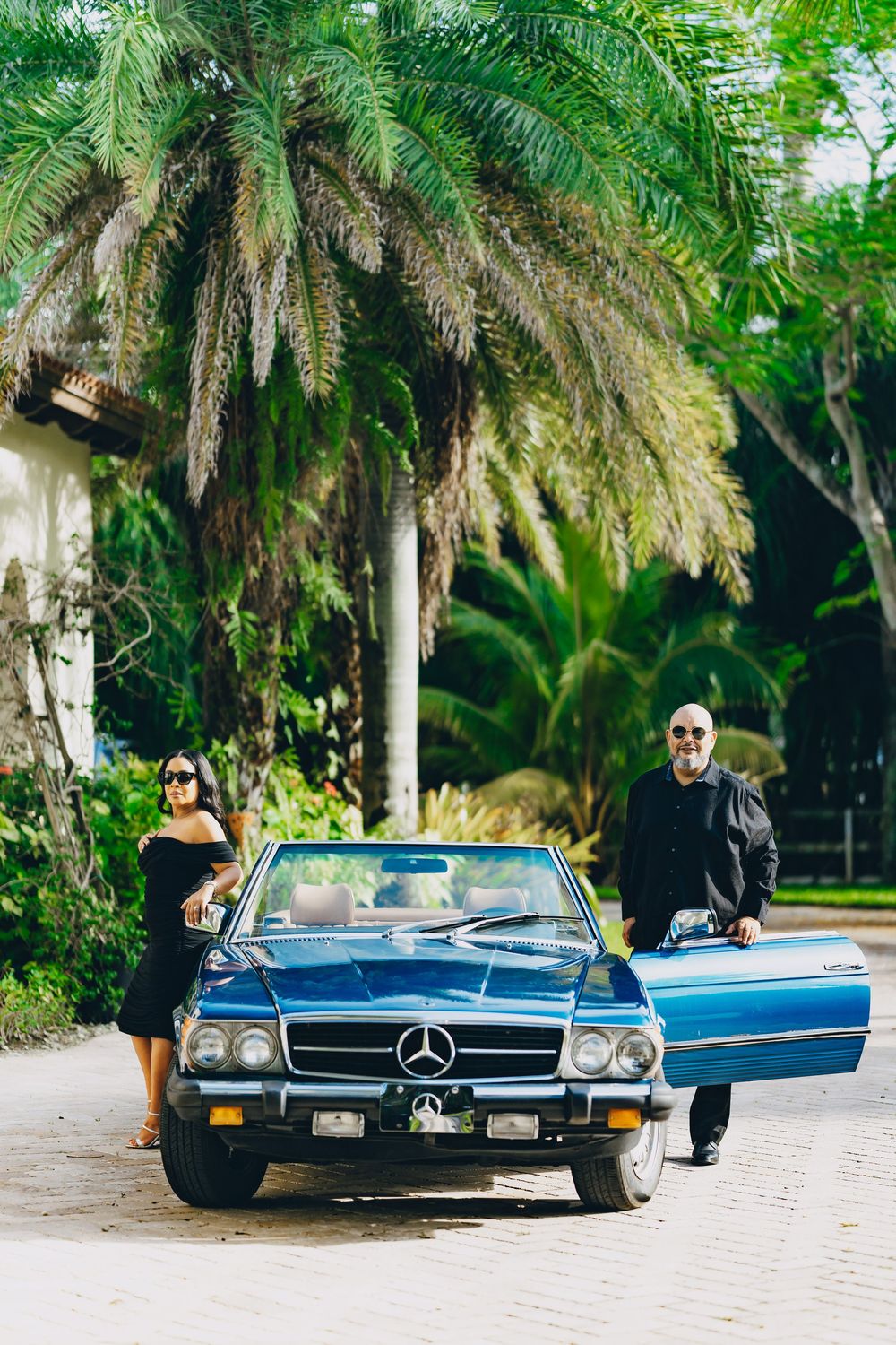 A vintage blue Mercedes-Benz convertible parked on a palm-lined driveway in a tropical setting.