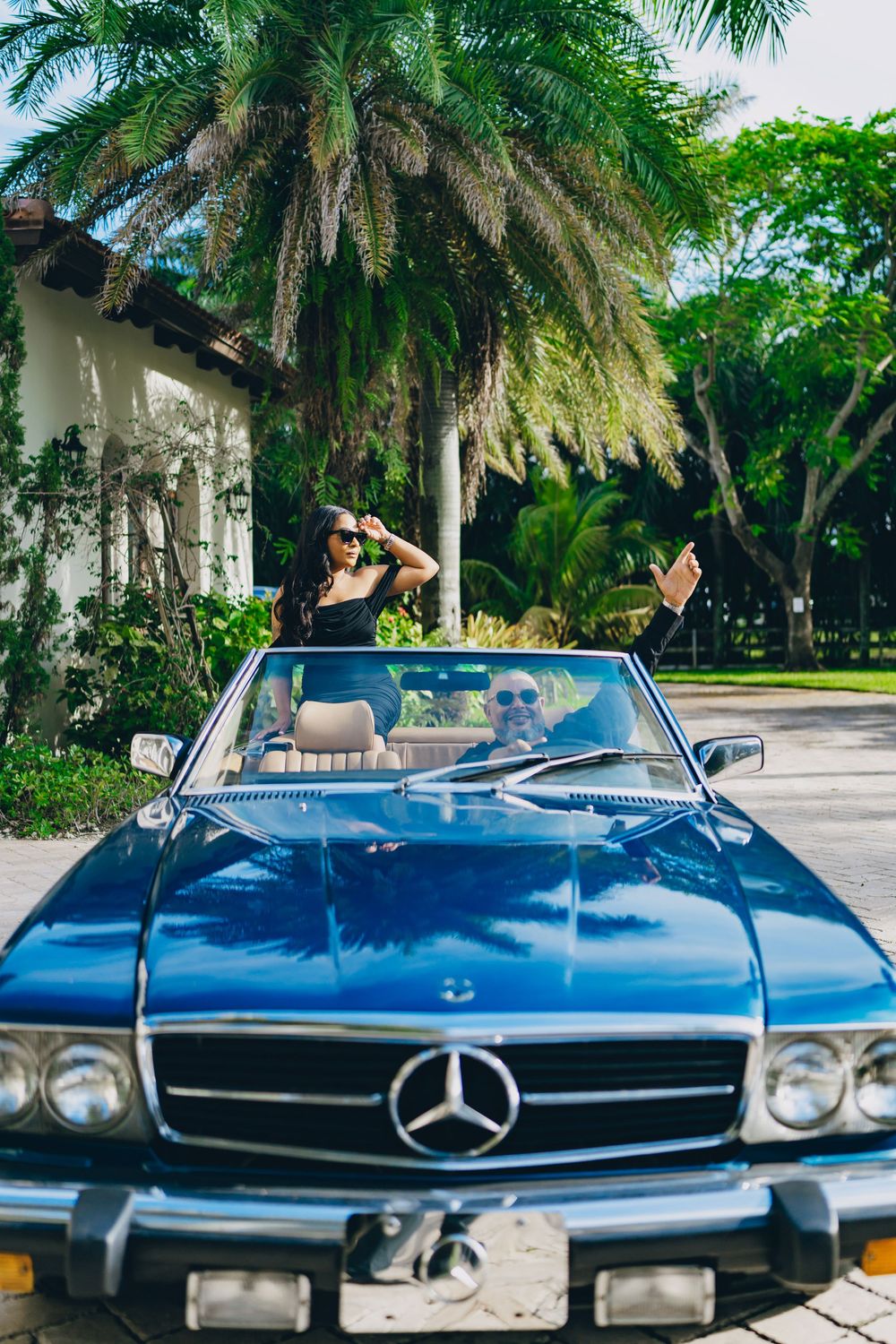 Classic blue Mercedes-Benz convertible parked under swaying palm trees in a tropical driveway.
