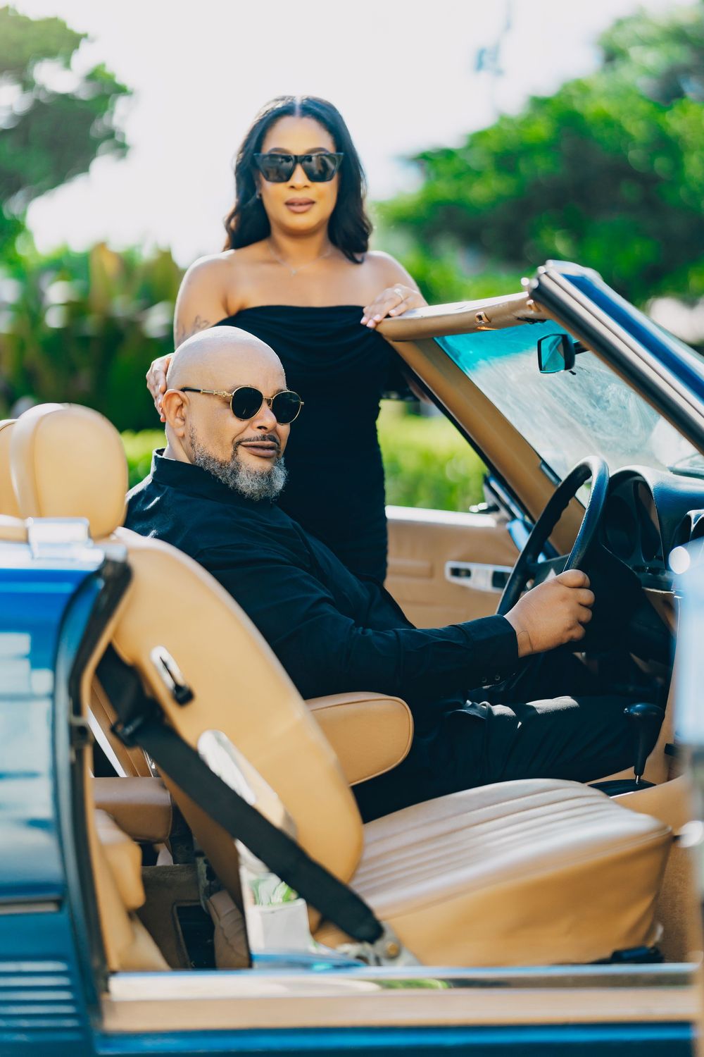 A stylish couple wearing sunglasses enjoy a sunny day in a classic convertible car with tan leather seats.
