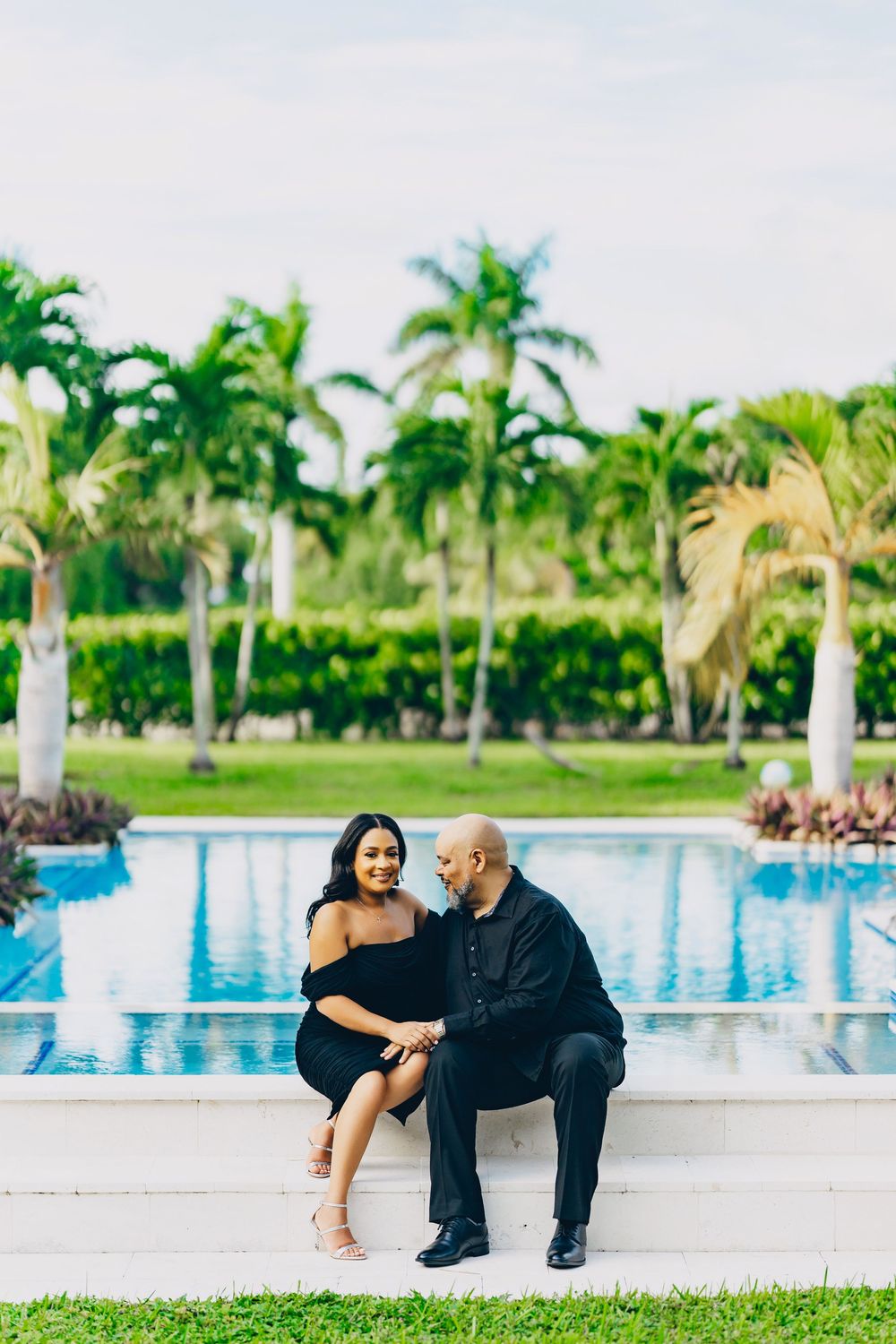 A couple sits together by a tropical resort swimming pool surrounded by palm trees on a sunny day.
