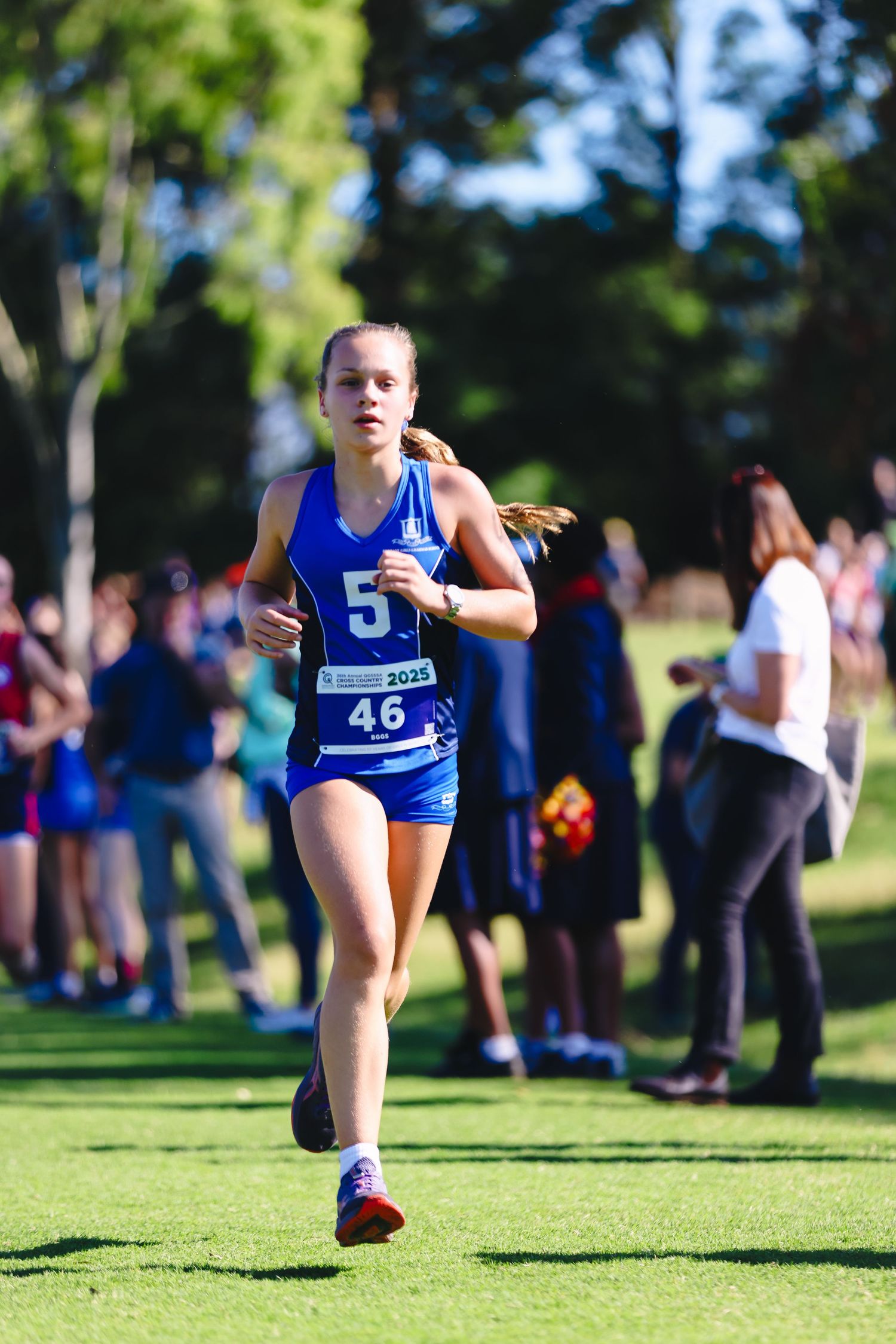 Runner in blue racing gear sprints across finish line at outdoor track competition with spectators and tents visible.