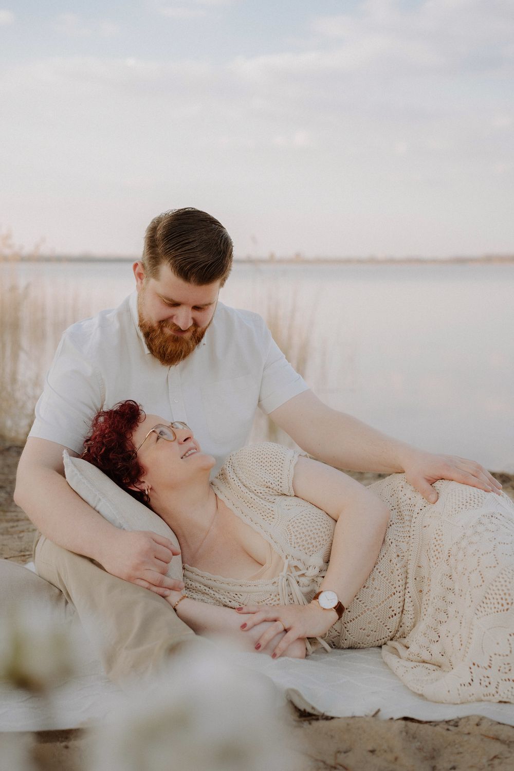A couple shares an intimate moment on a blanket by a serene lake with tall grass and soft cloudy skies in the background.
