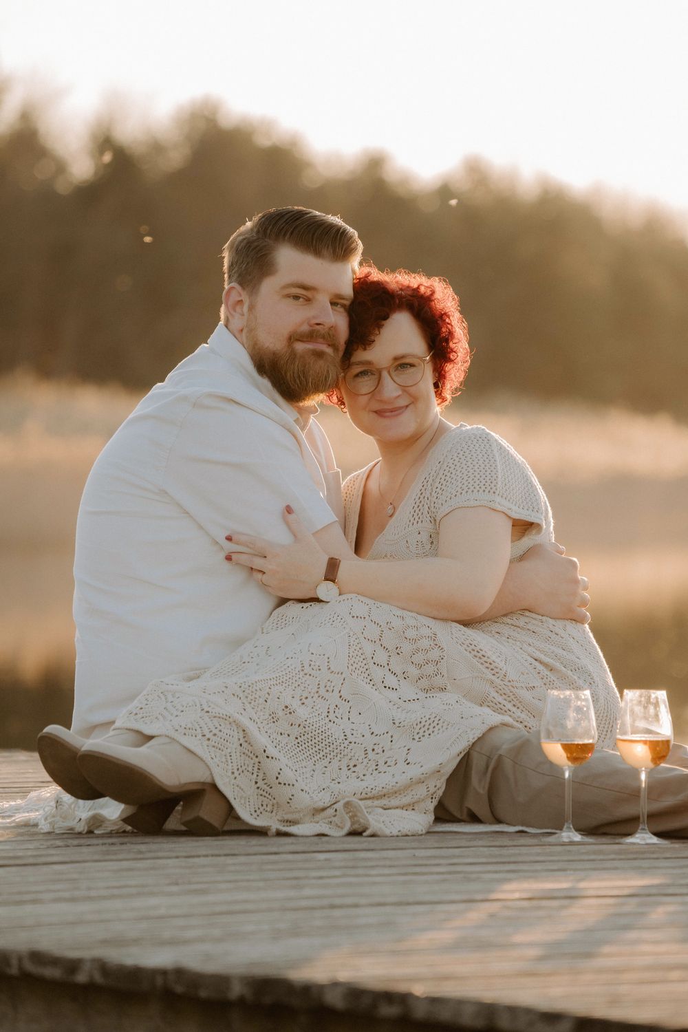 Romantic couple sharing intimate moment at sunset on dock with wine glasses beside them in dreamy lighting.