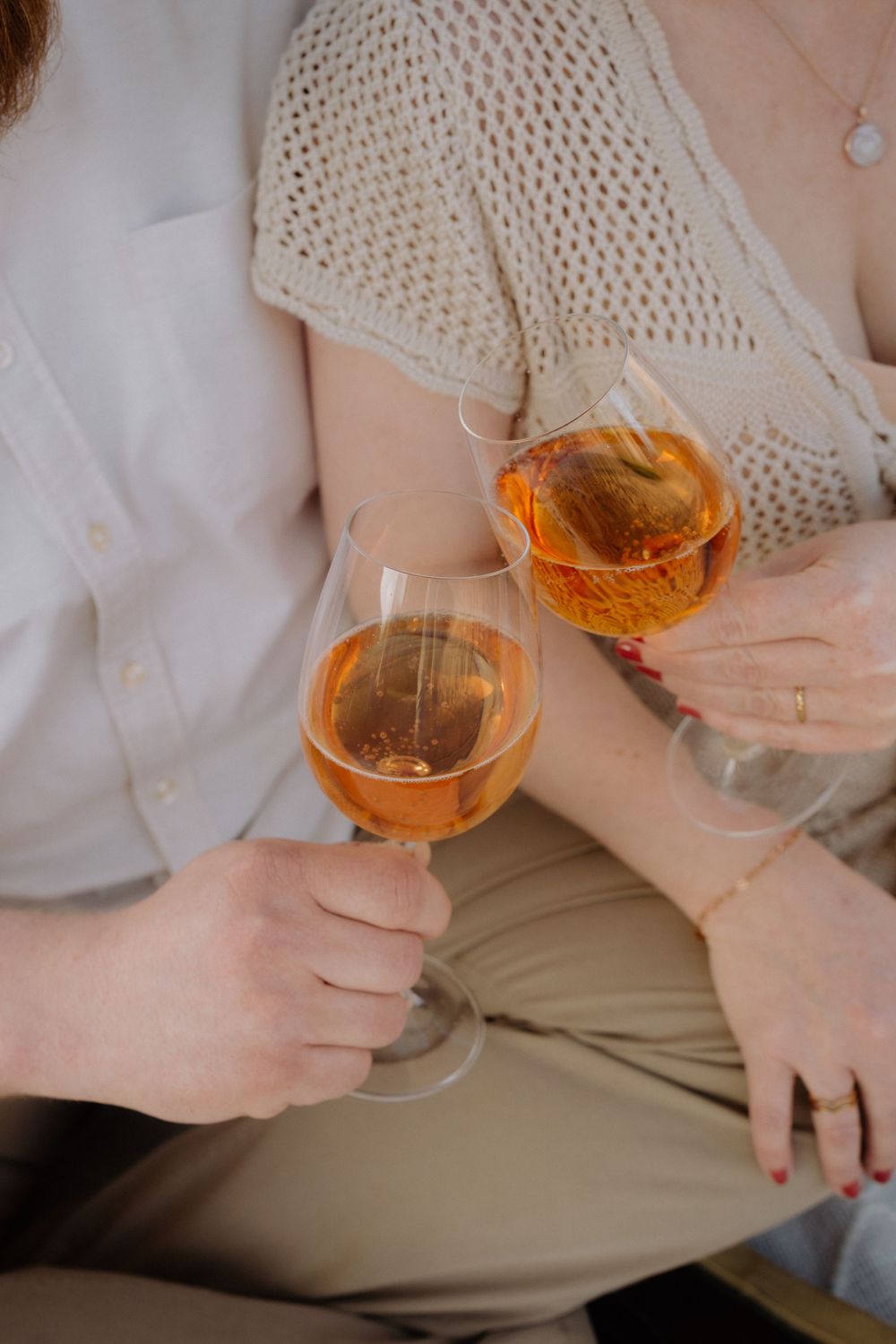 Two people in casual attire toasting with glasses of rose wine, wearing cream knit top and beige pants.