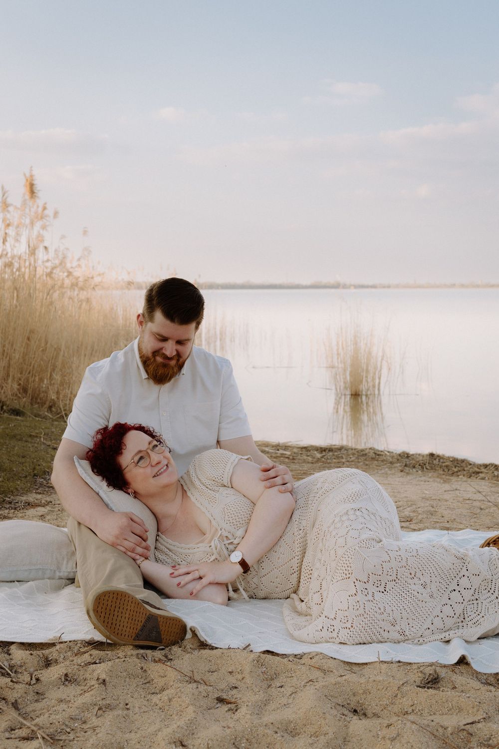 Couple relaxes on blanket by lakeside at sunset with tall reeds and calm water in background.