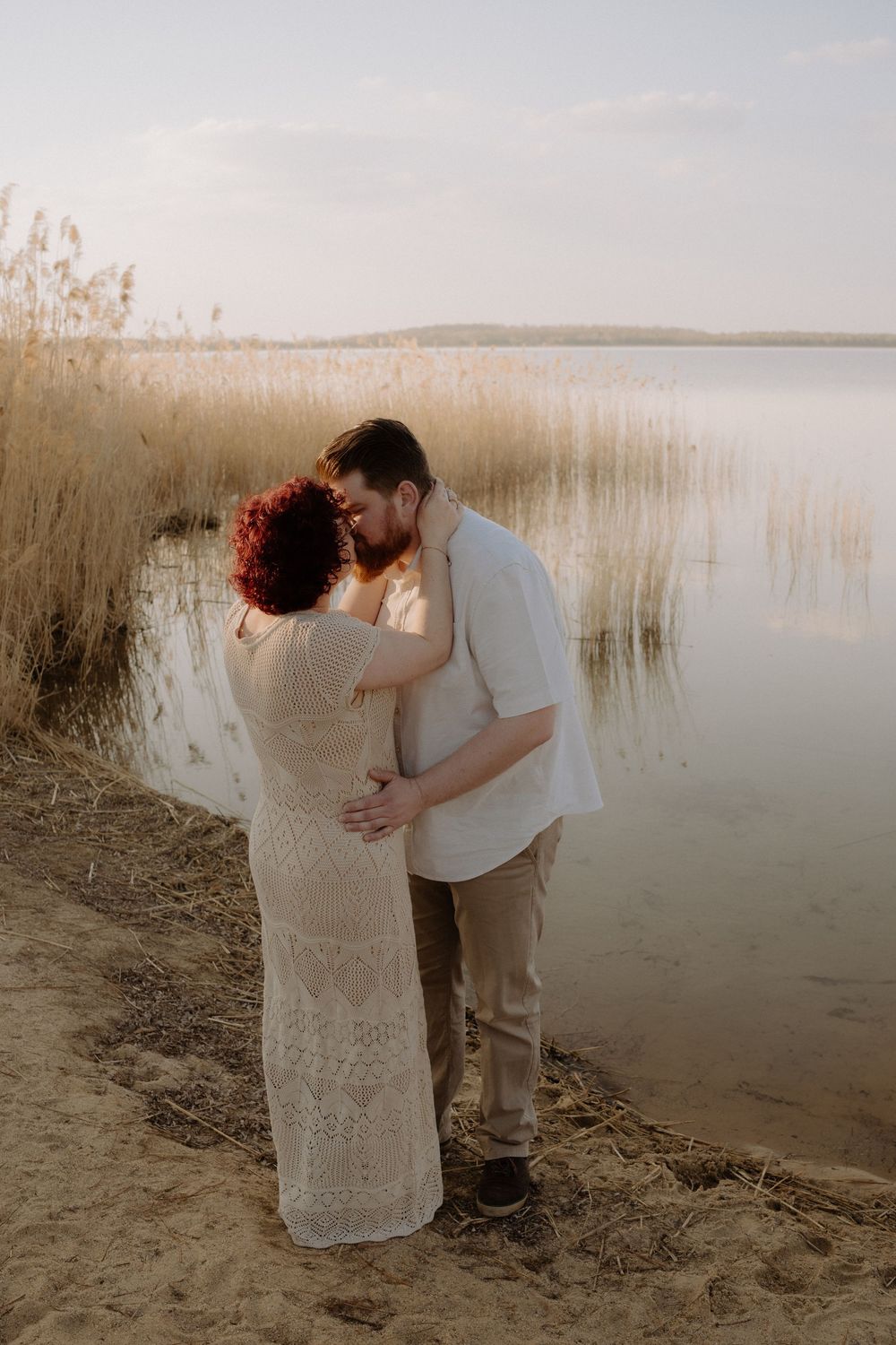 A romantic couple embraces at sunset by a serene lake surrounded by tall reeds and golden light.