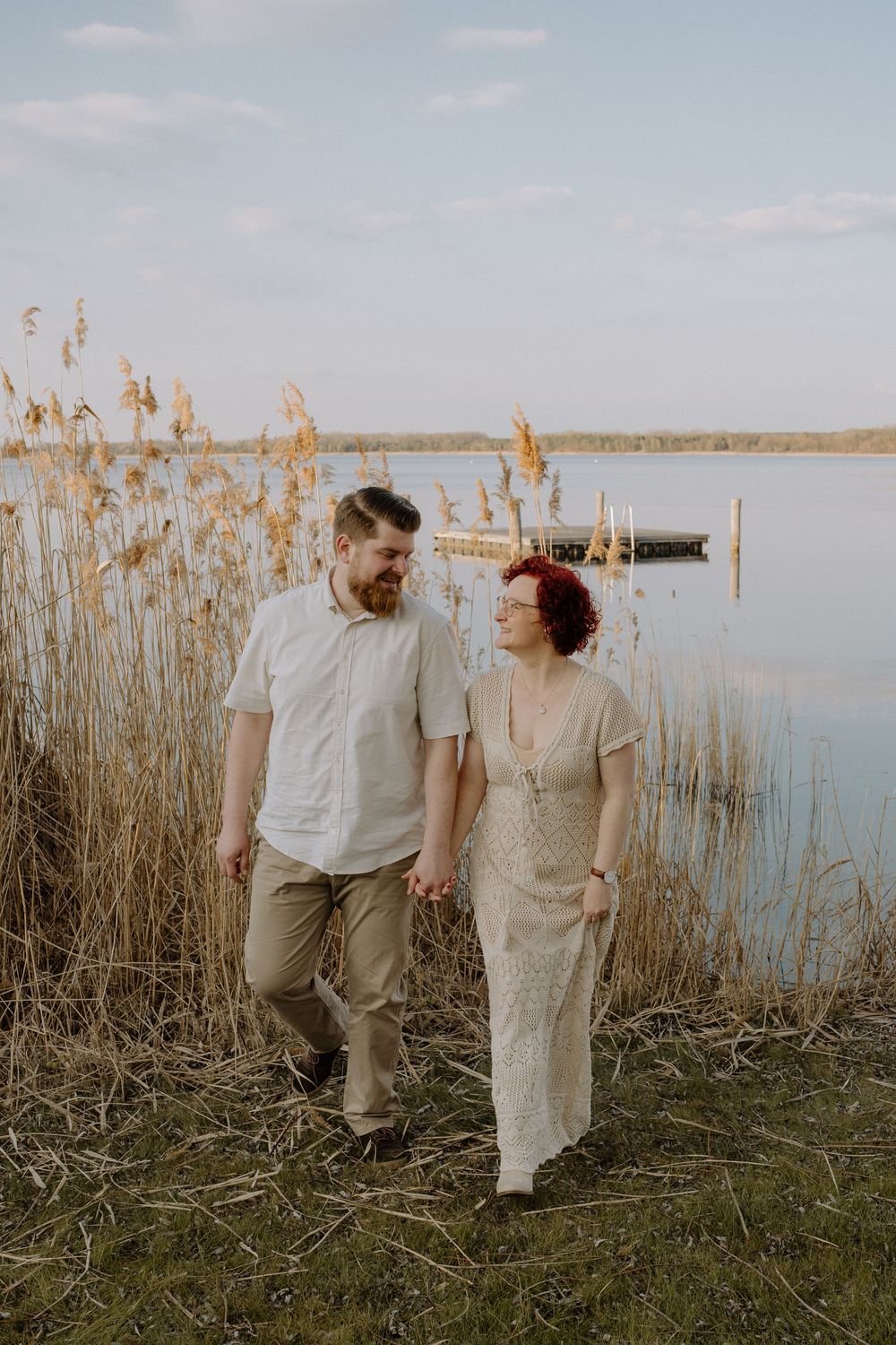A couple walks hand in hand along a lakeside path with tall reeds and a boat visible in the serene water at sunset.
