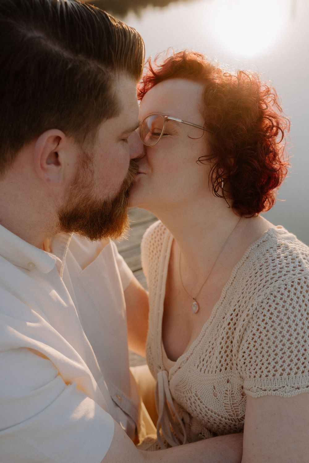 A romantic couple shares a tender kiss in warm sunlight while wearing white and cream colored clothing.