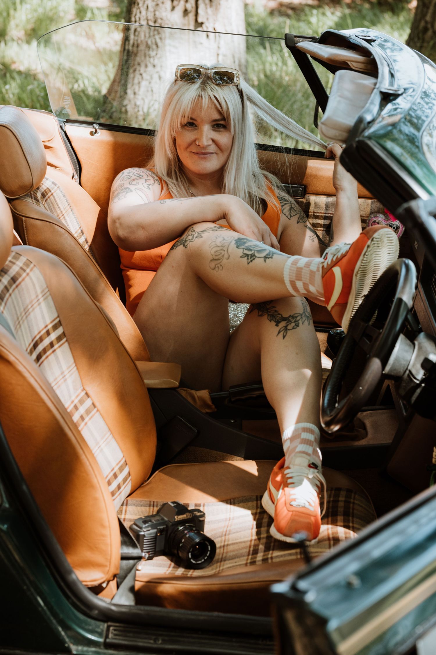 Vintage car interior with plaid tan leather seats and a camera visible on the passenger seat on a sunny day.